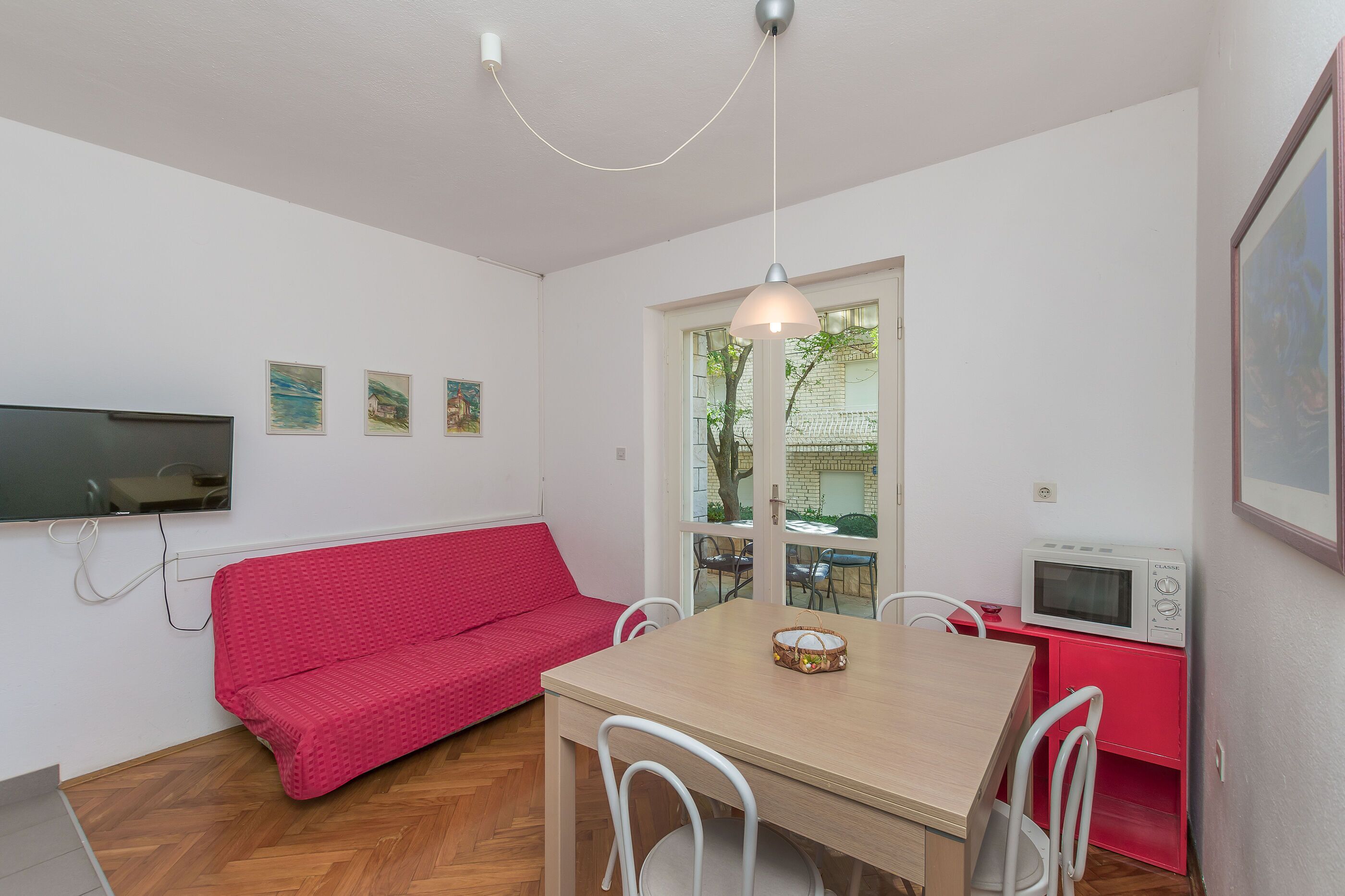 Ein kompakter Wohnraum mit einem roten Sofa, einem Holzesstisch, weißen Stühlen, einem an der Wand montierten Fernseher und einer Mikrowelle auf einem roten Schrank.