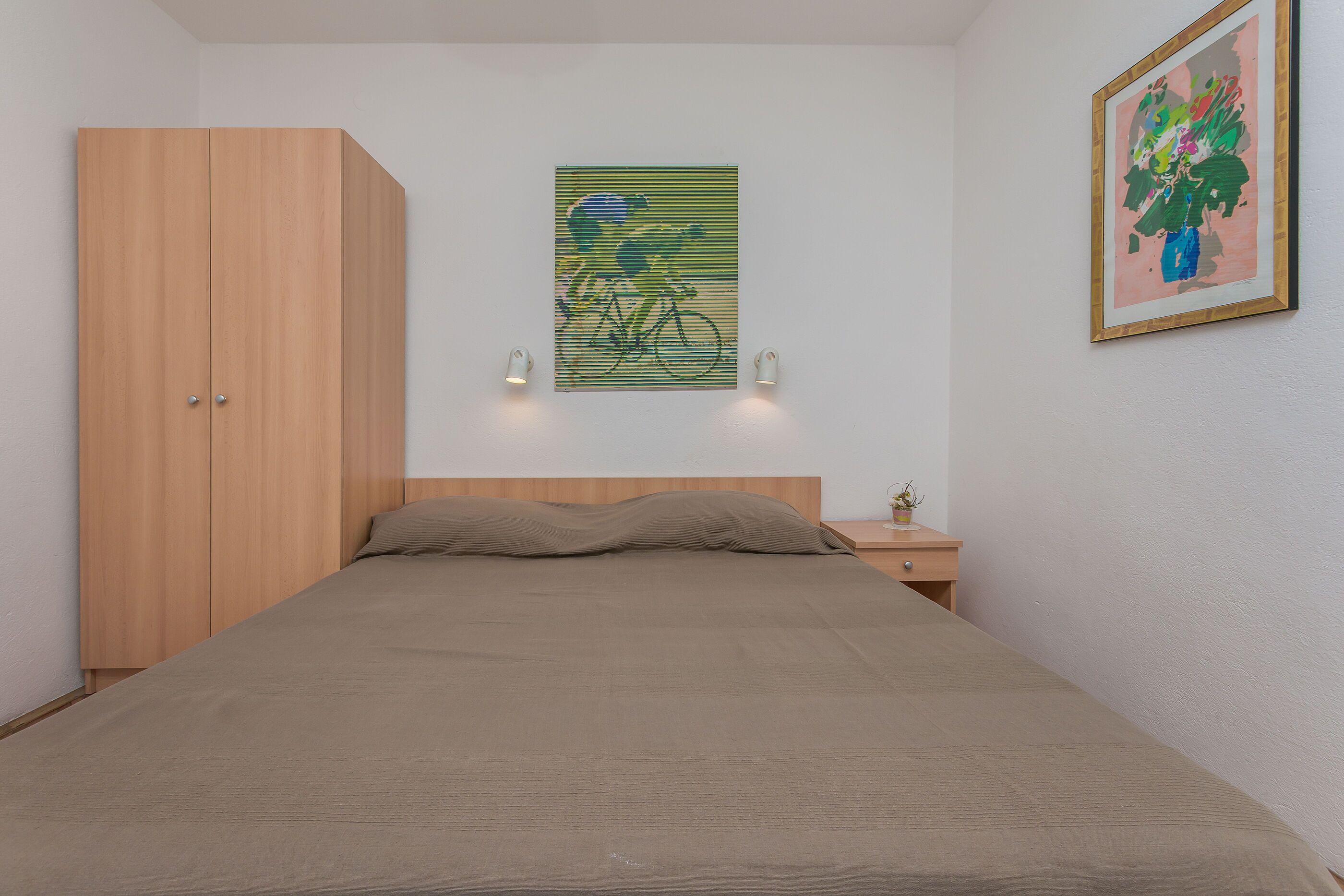 A bedroom with a bed, wooden wardrobe, two wall-mounted lights, and two framed artworks.