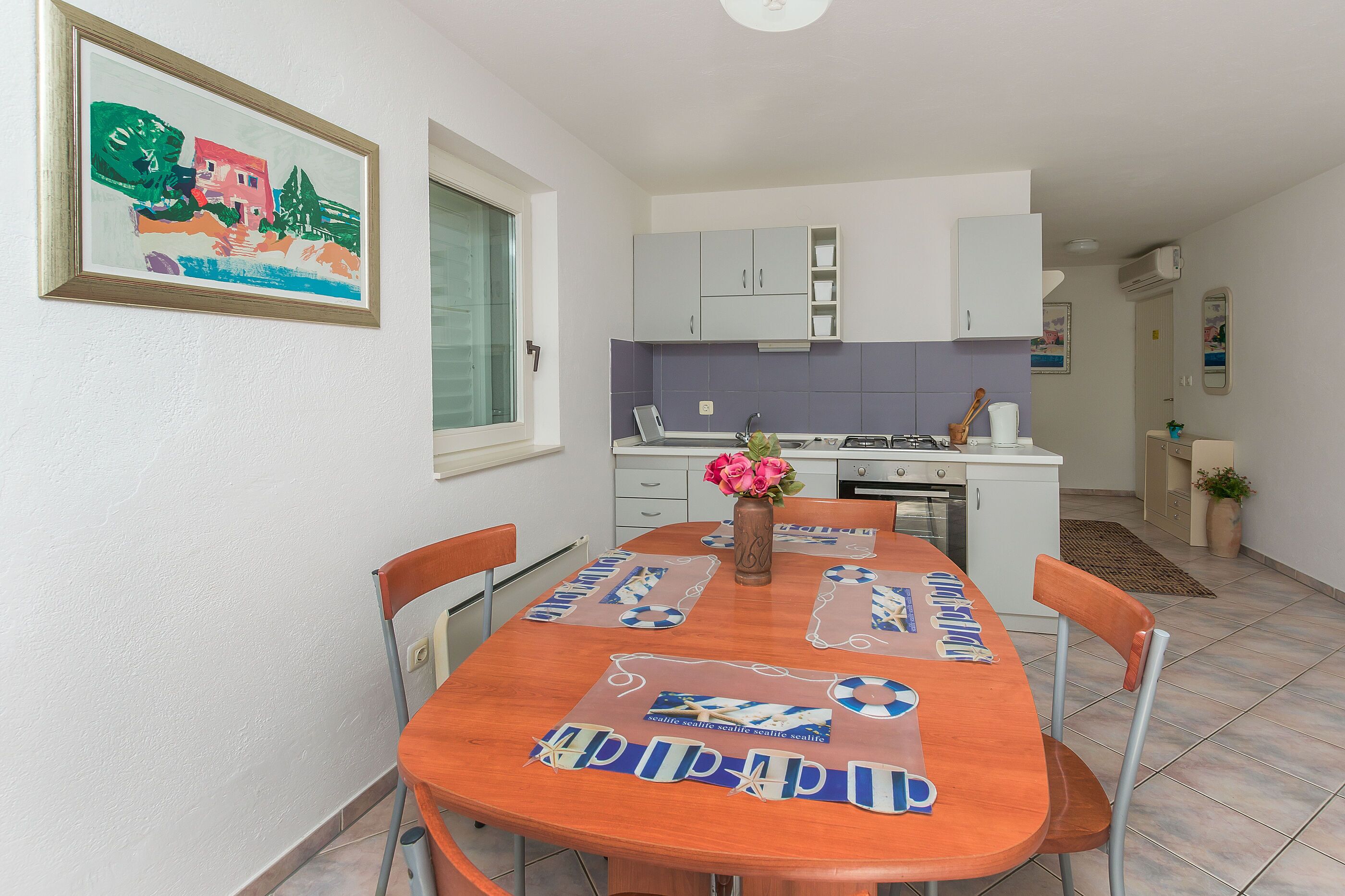 A dining area with a wooden table, chairs, and a vase with flowers. There is a kitchen in the background with cabinets and a stove.
