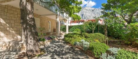A paved pathway leading to a building with a stone facade, surrounded by lush greenery and a mountain backdrop.