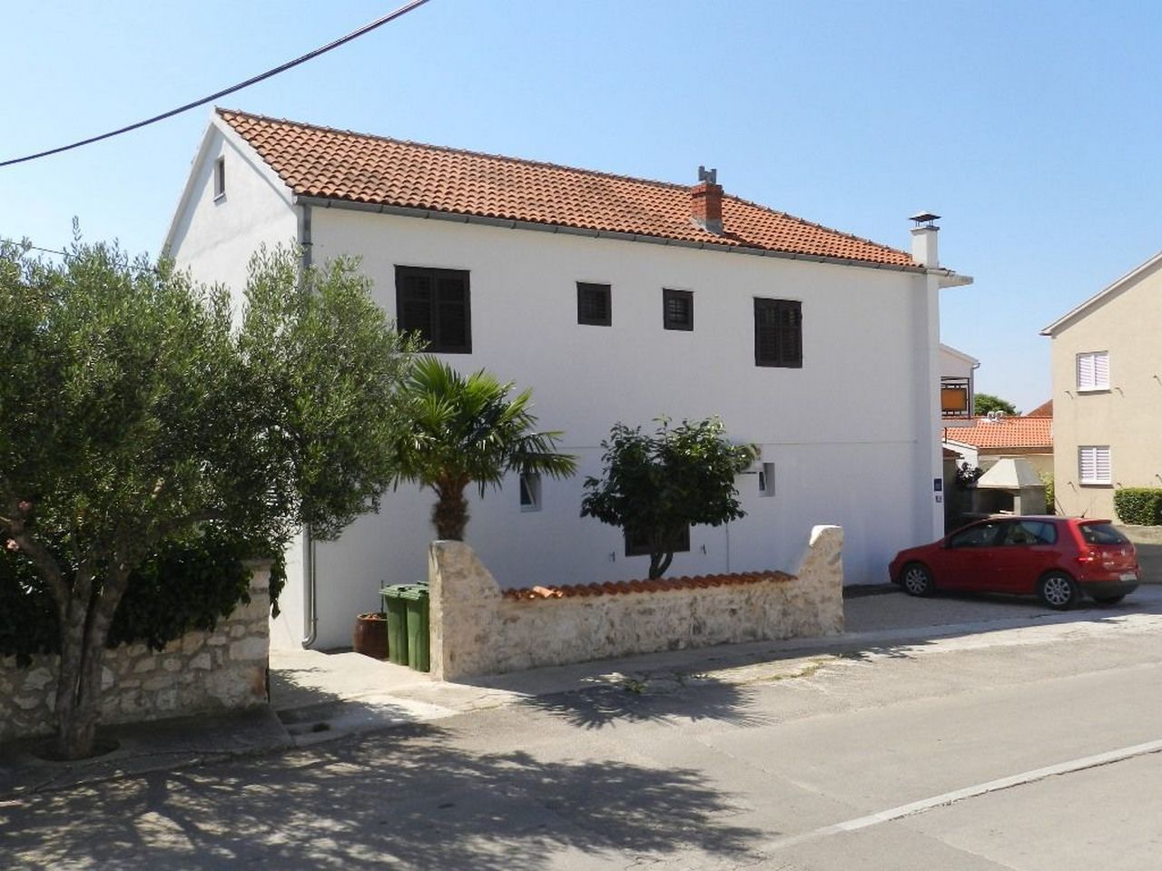 Un edifici blanc amb teules vermelles al terrat, un arbre petit i un cotxe vermell estacionat a la façana.