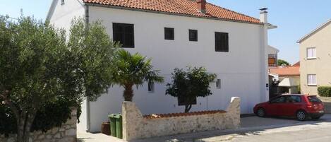 A white building with a red-tiled roof, a small tree, and a red car parked in front.