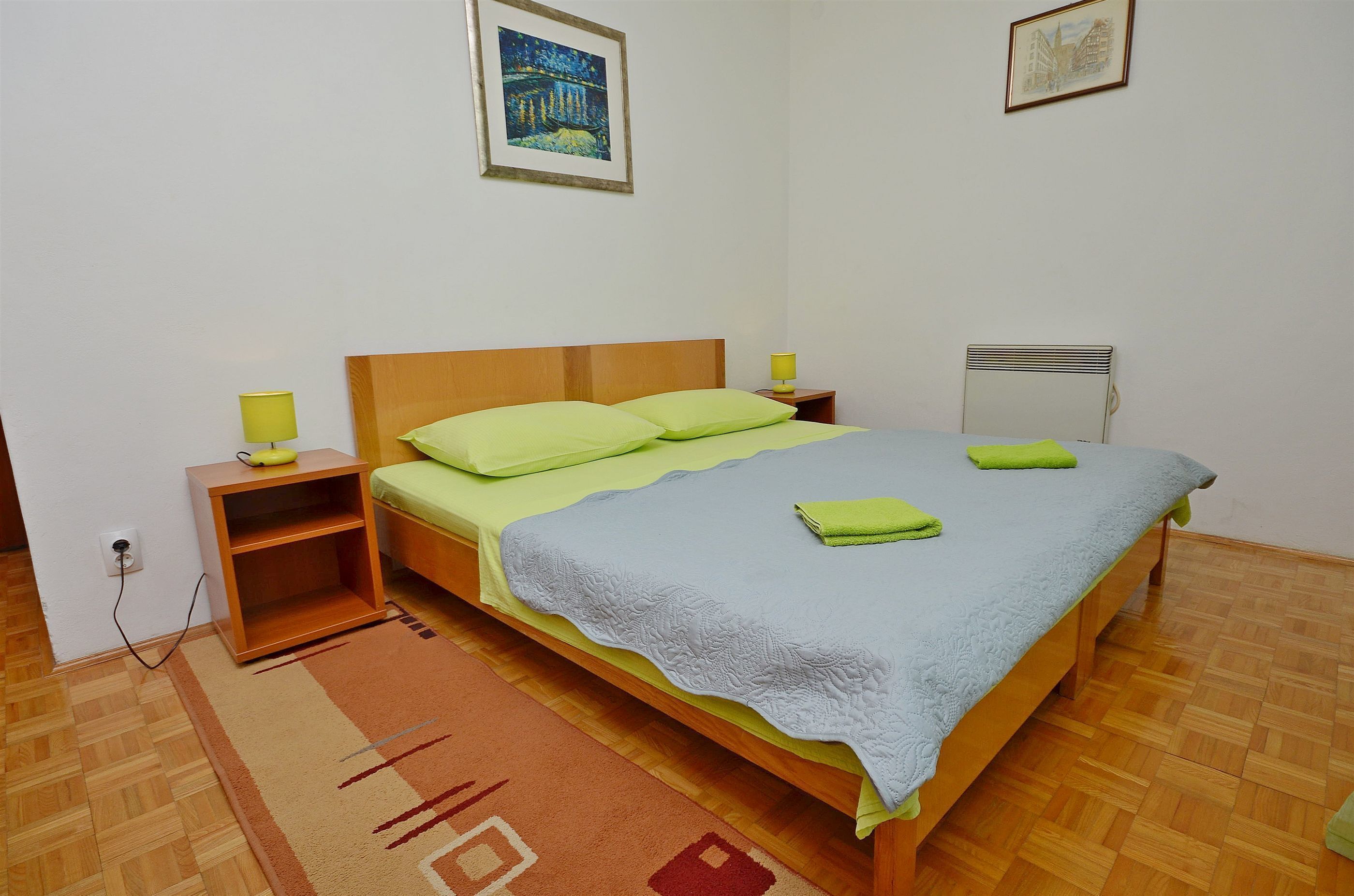 Una cama bien tendida con almohadas verdes y una manta azul claro, una mesita de noche de madera con una lámpara y un radiador en la pared.