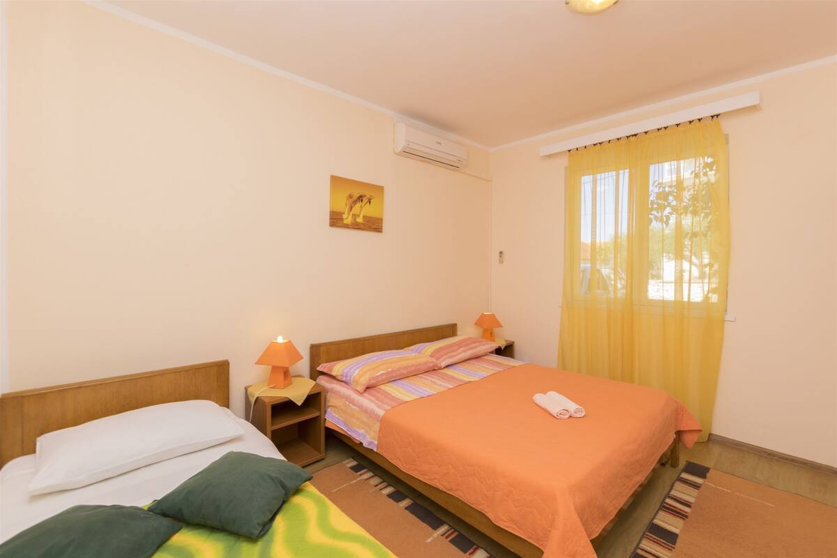 A hotel room with two beds, a window with yellow curtains, an air conditioner, and a small painting on the wall.