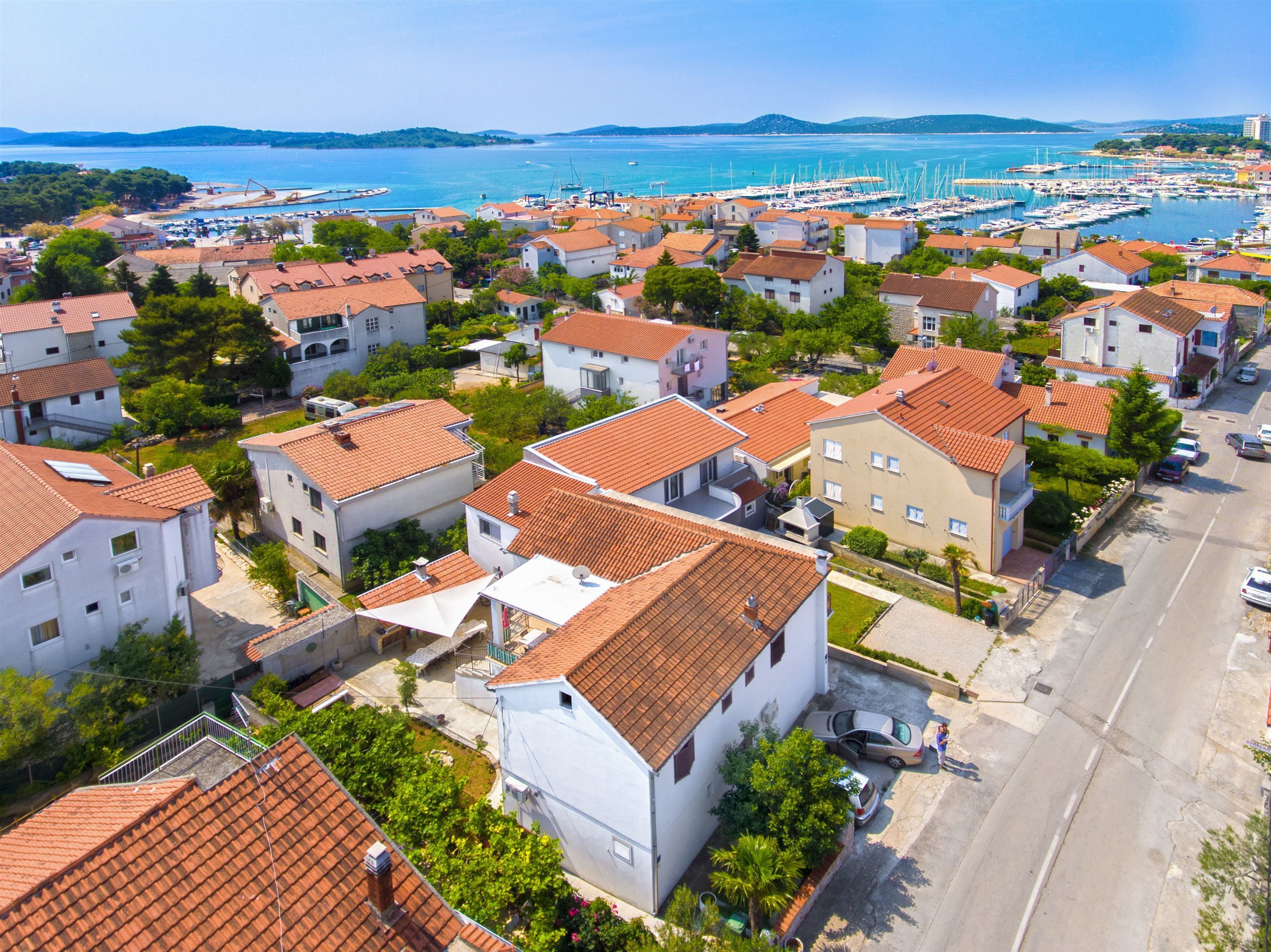 Un área residencial costera con casas de techos de teja anaranjada, un puerto deportivo con barcos y una vista despejada del mar.