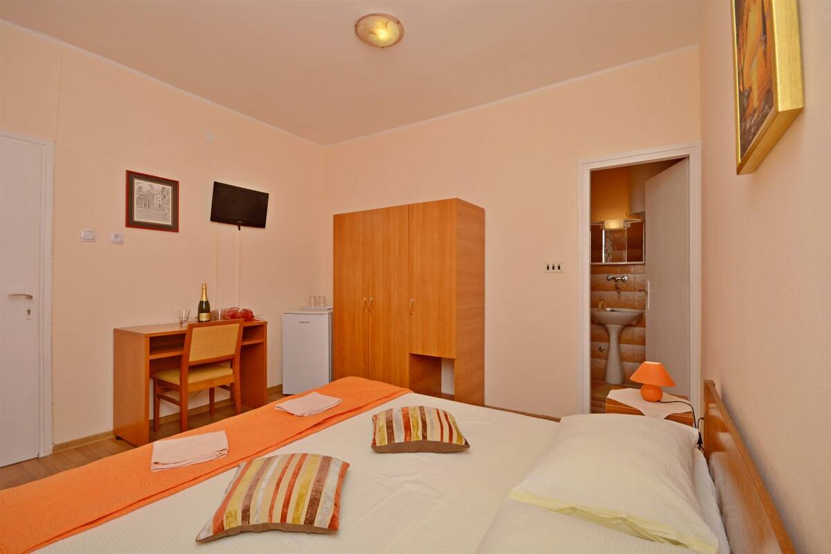 A hotel room with a bed, a desk, a wardrobe, a television, and a bathroom visible through an open door.