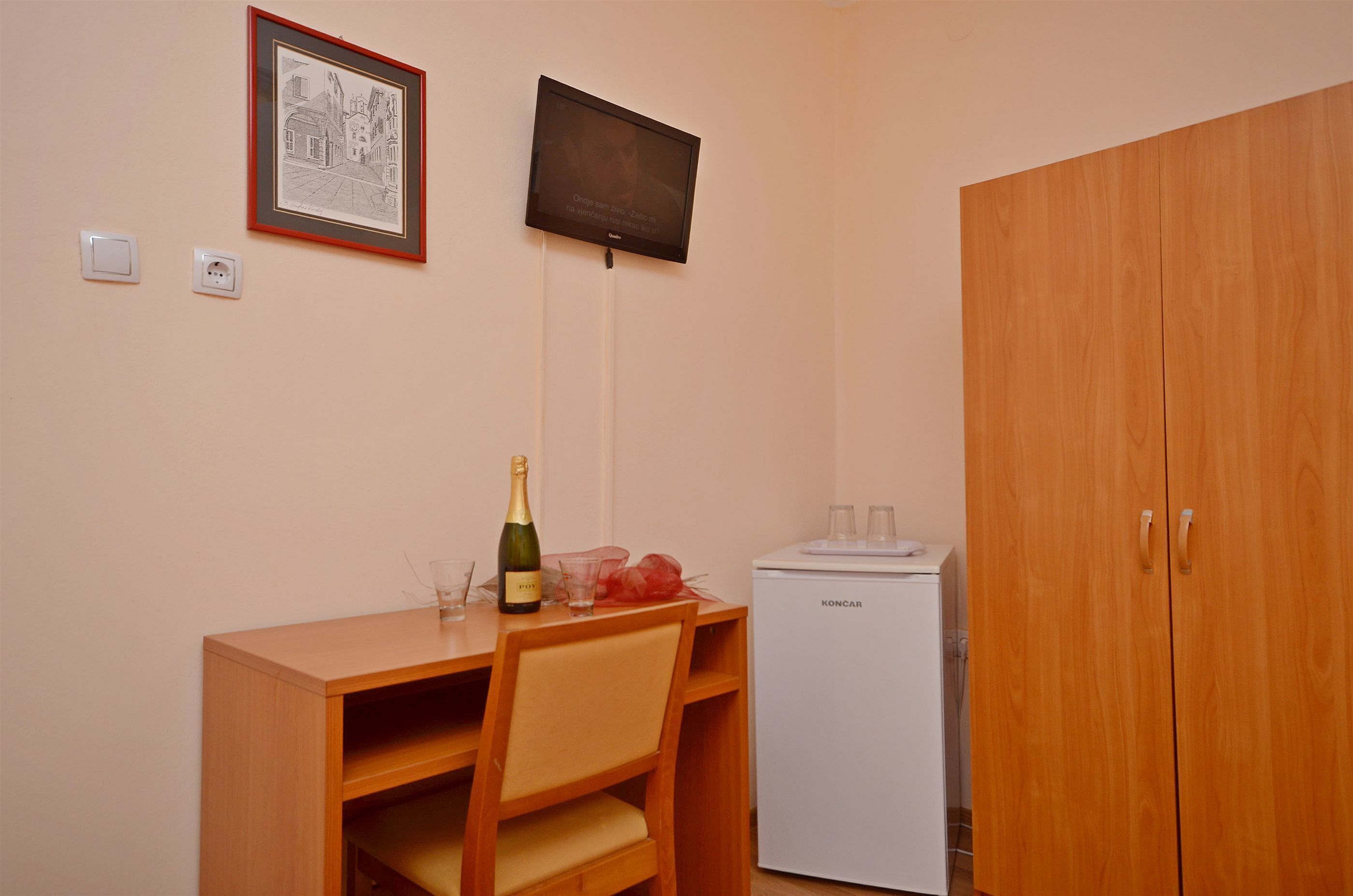 A room with a wooden desk, a chair, a bottle of champagne, a small refrigerator, and a wardrobe.