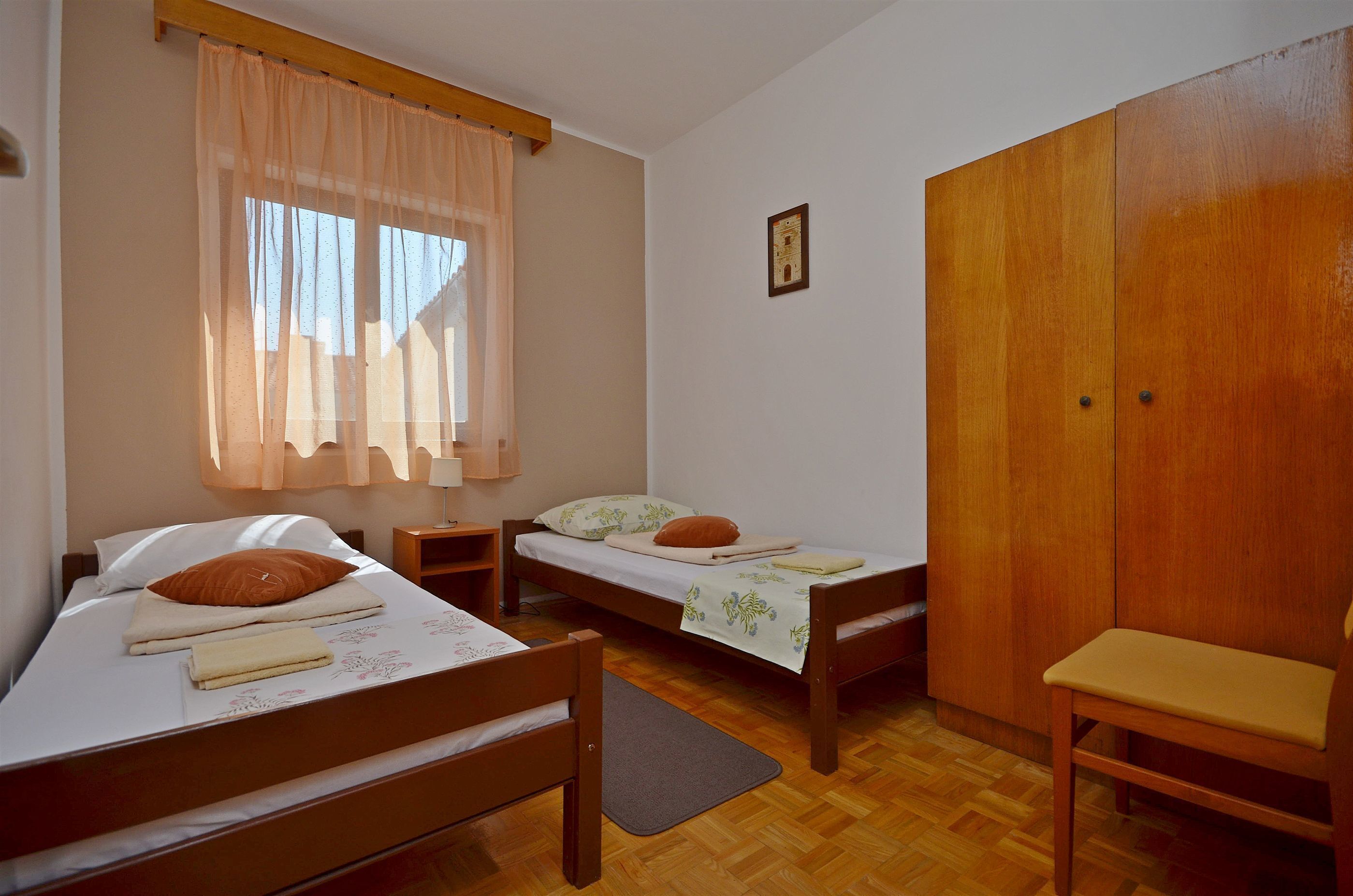 A room with two wooden beds, a wooden wardrobe, a wooden chair, and a window with sheer curtains.