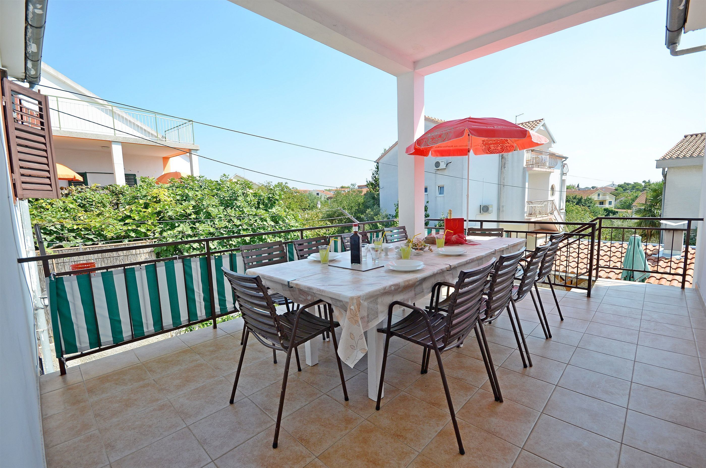 Ein Balkon mit Tisch und Stühlen, einem roten Sonnenschirm und Blick auf Grünflächen und Gebäude.