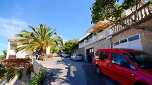 A street with parked cars, a red car on the right, and a building with a stone facade and a balcony. - Apartments Jakov (Jelsa)