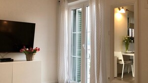 A bedroom with a TV on a white cabinet, a bed with a floral bedspread, a window with curtains, and a door leading to a bathroom. - Apartments Mirko (Seget Vranjica)