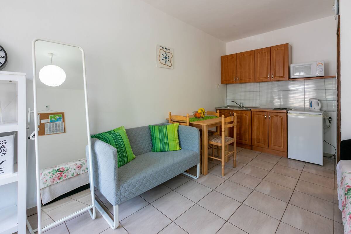 Un espacio habitable compacto con kitchenette, un sofá con cojines verdes, una mesa de comedor y un refrigerador pequeño.
