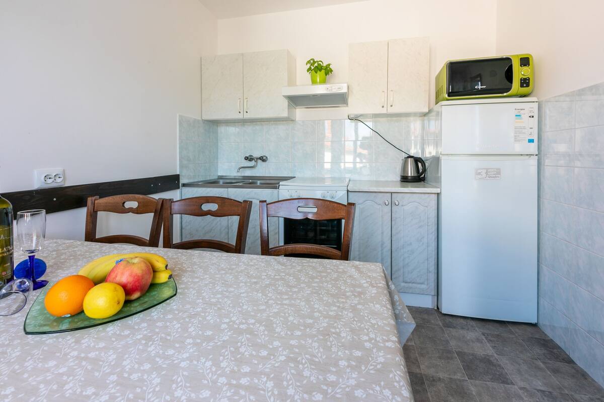 Una cocina con una mesa de comedor, sillas, un frutero, microondas y refrigerador.