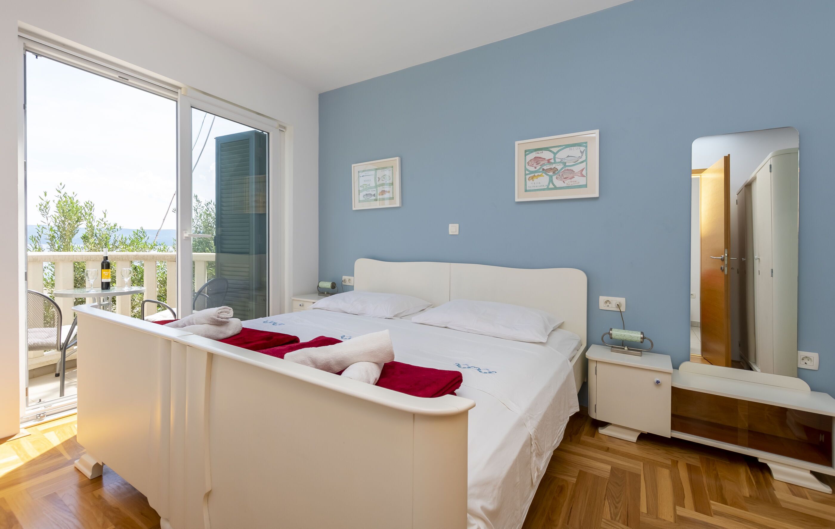 A bedroom with a bed, a bedside table, a nightstand, a mirror, and a window with a balcony.