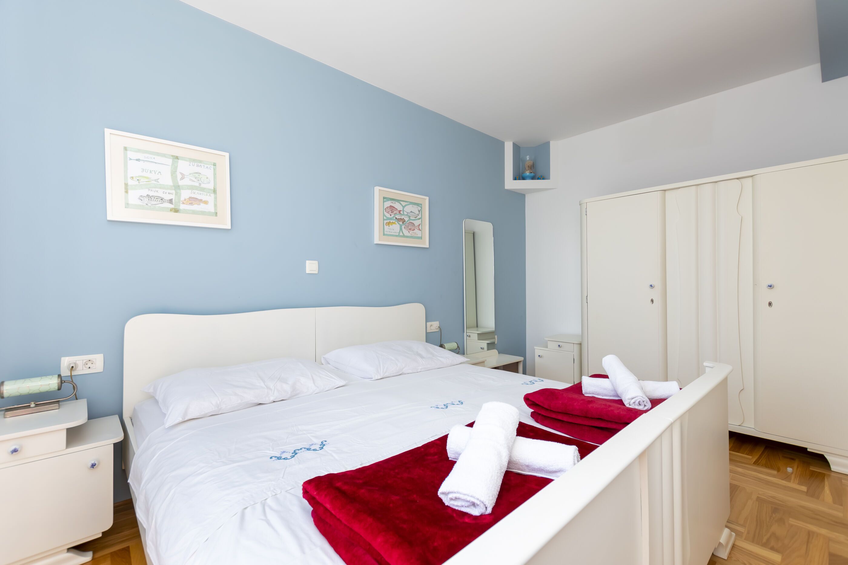 A neatly made bed with white linens and a red blanket, a bedside table with a lamp, a wall-mounted mirror, and a wardrobe with a mirror.
