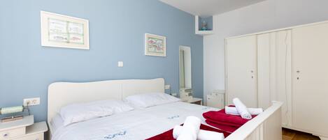 A neatly made bed with white linens and a red blanket, a bedside table with a lamp, a wall-mounted mirror, and a wardrobe with a mirror.