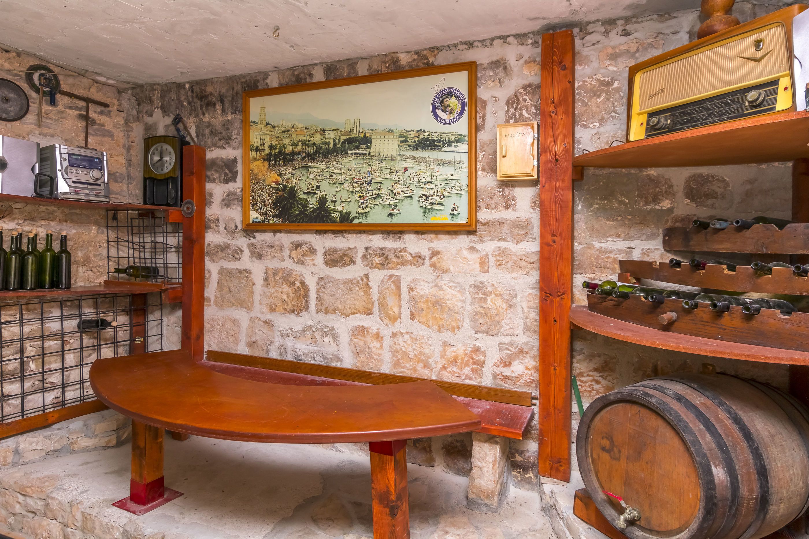 A rustic room with a stone wall, a wooden table, wine bottles, a vintage radio, and a large framed picture.