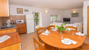 Cocina compacta con estufa, microondas y refrigerador. Cuenta con una zona de comedor con mesa y sillas de madera, y un televisor al fondo.