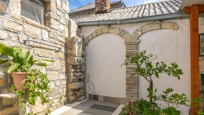 Un edificio de piedra con techo de tejas, una puerta blanca y un pequeño jardín con macetas y flores.