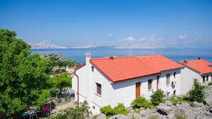 A white building with a red roof, surrounded by greenery and rocky terrain, overlooking a body of water and distant mountains.