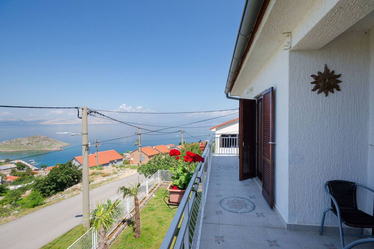 A balcony with a view of the sea and houses, a chair, and a decorative sun on the wall.