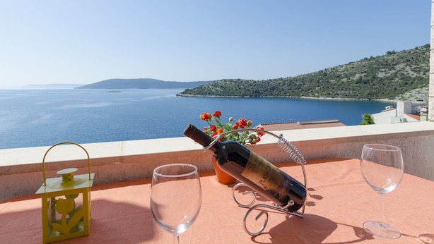 Une table avec une bouteille de vin, des verres et une lanterne, placée sur un balcon donnant sur la mer et un paysage vallonné.