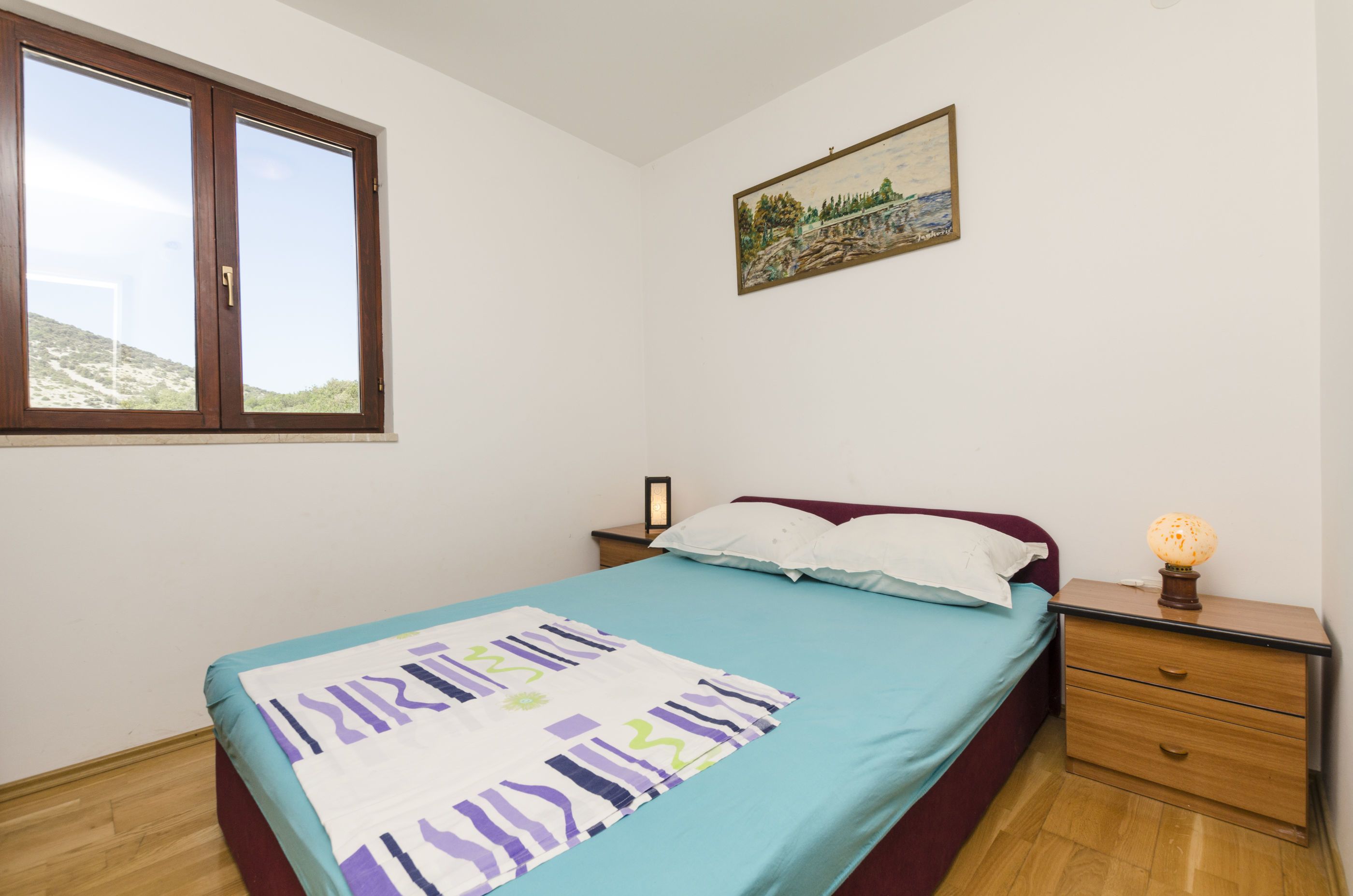Un dormitorio con cama, mesita de noche, lámpara, un cuadro en la pared y ventana.