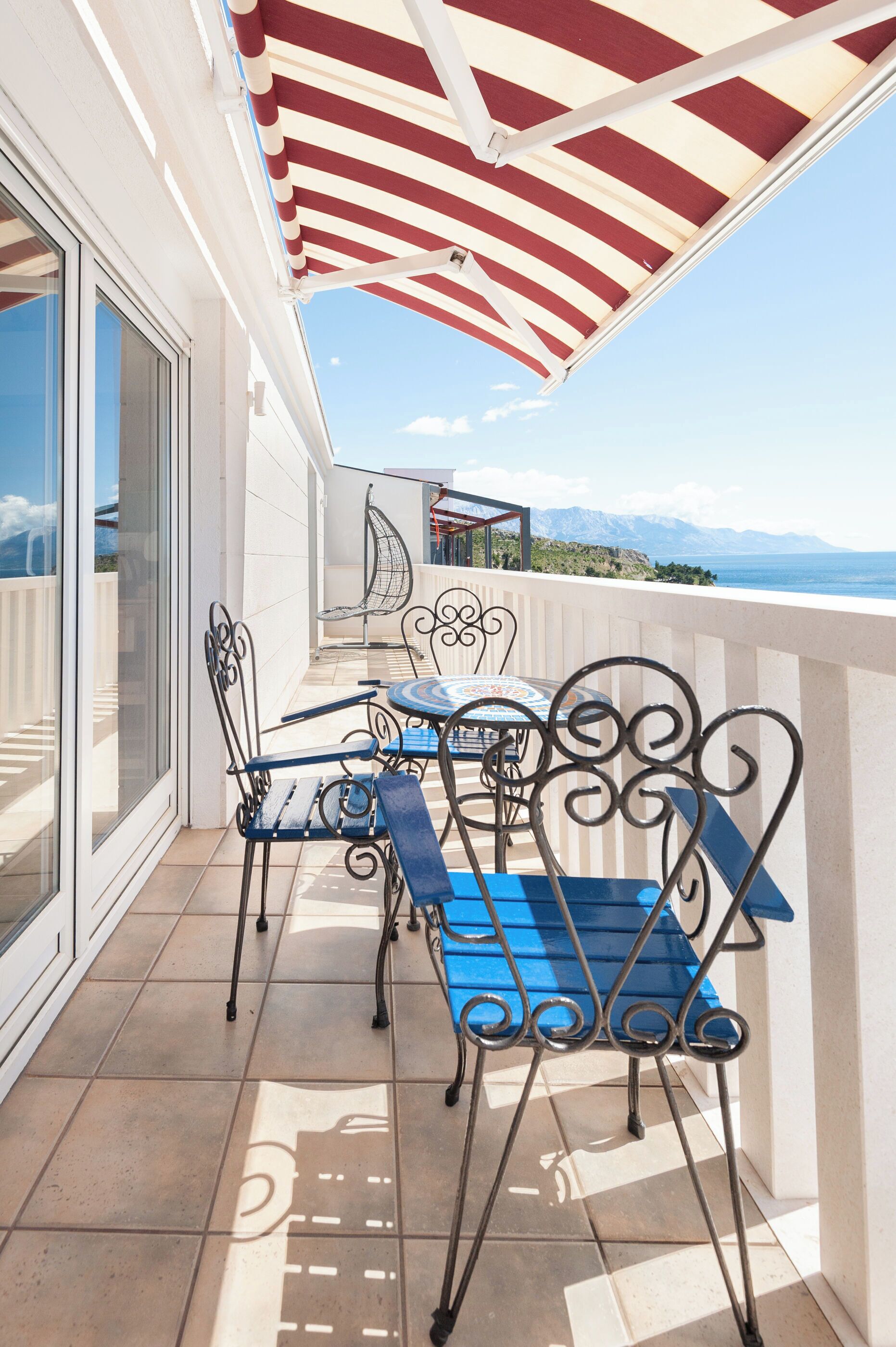 A balcony with a striped awning, metal chairs, a table, and a view of the sea and mountains.