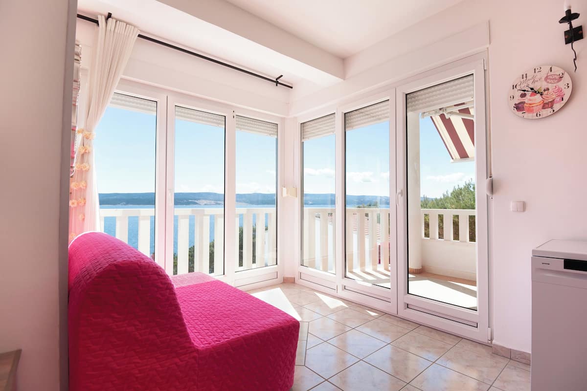 A balcony with a view of the sea, a pink chair, and a white wall with a clock.