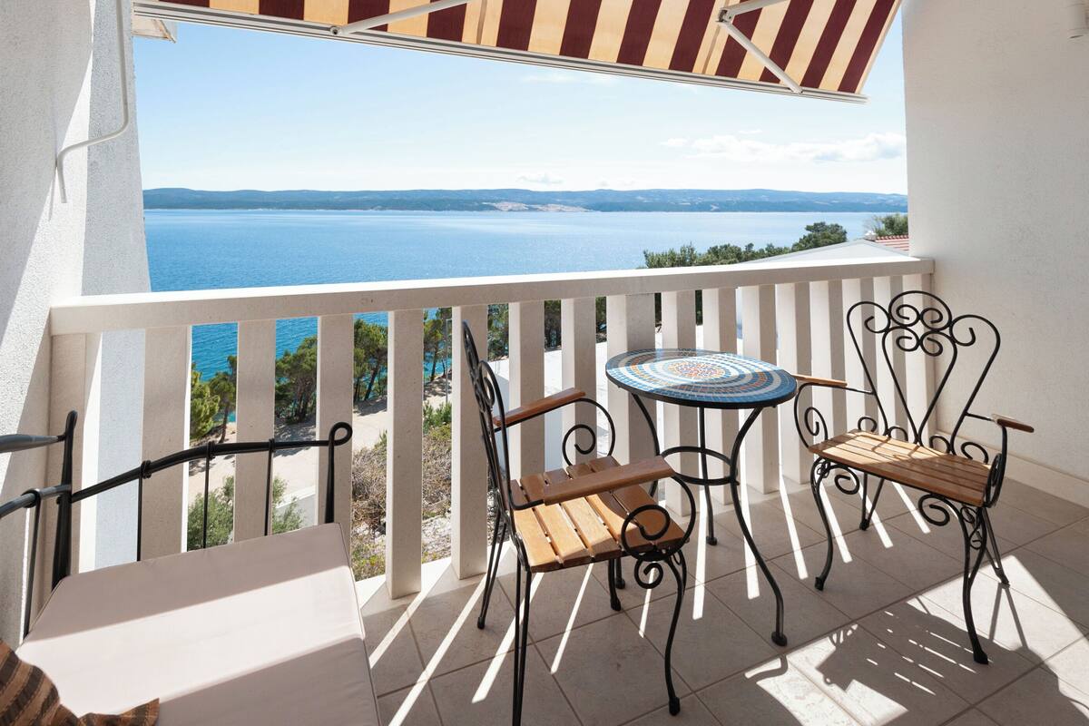 A balcony with a view of the sea, two wooden chairs, a small round table, and a striped awning.