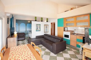 A compact living space with a dining area, a sofa, a kitchen with wooden cabinets, and a view of a balcony. - Apartments Gordana (Lokva Rogoznica)