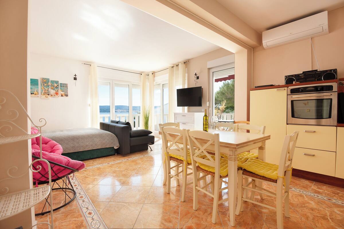 A compact living space with a bed, a dining area with chairs, a TV mounted on the wall, and an air conditioning unit.