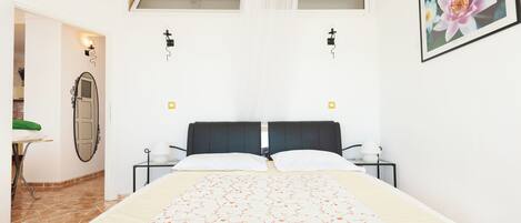 A bedroom with a bed, a nightstand, a wall-mounted light, a framed picture, and a window with a curtain.