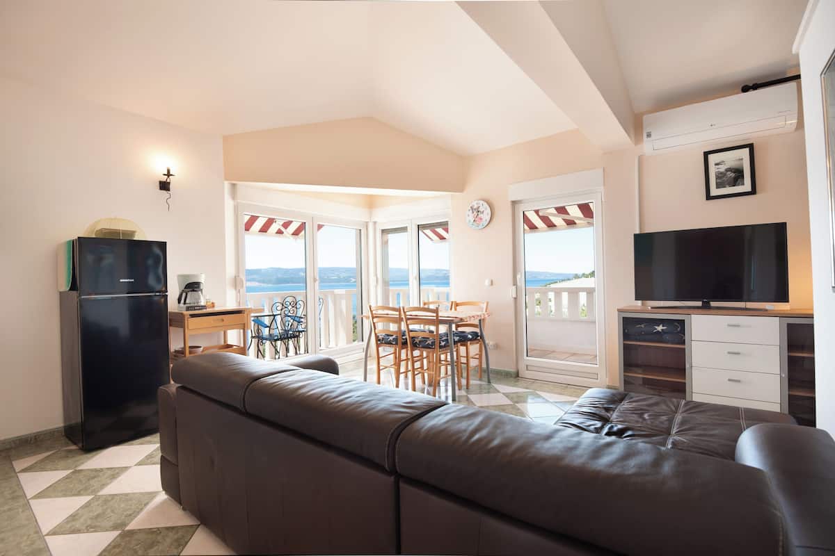 A living room with a sofa, a TV, a dining table with chairs, a refrigerator, and a view of the sea.