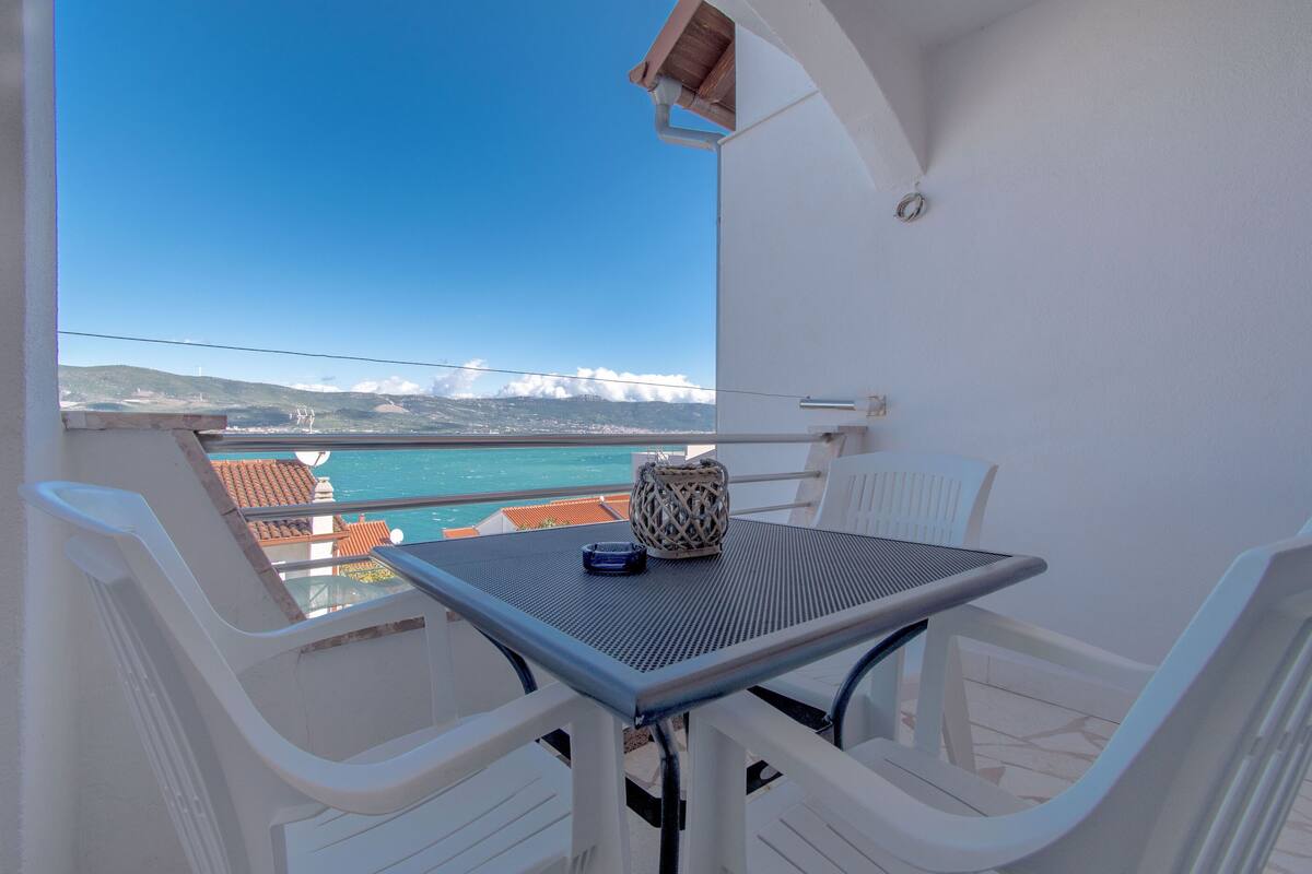 Un balcon avec une table et des chaises, offrant une vue sur la côte, avec des bâtiments et des montagnes visibles au loin.