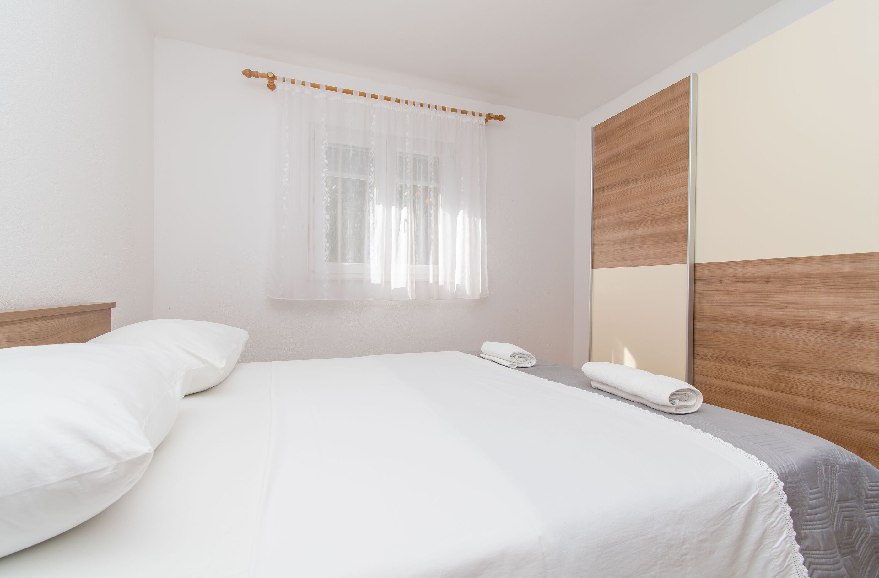 A neatly made bed with white linens, a wooden headboard, and a window with sheer curtains.