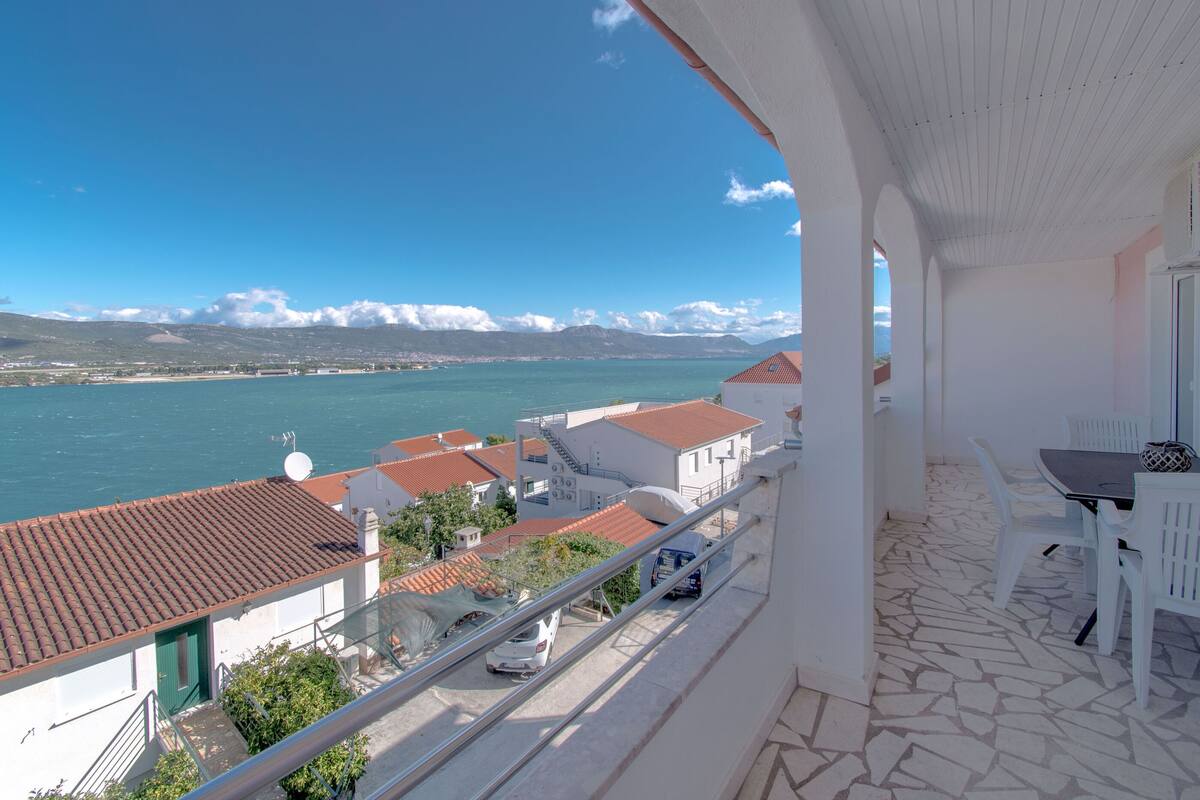 Un balcon donnant sur une ville côtière, un coin repas avec des chaises blanches et un sol carrelé.