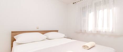 A neatly made bed with white linens and pillows, a rolled towel on the bed, and a window with sheer curtains.