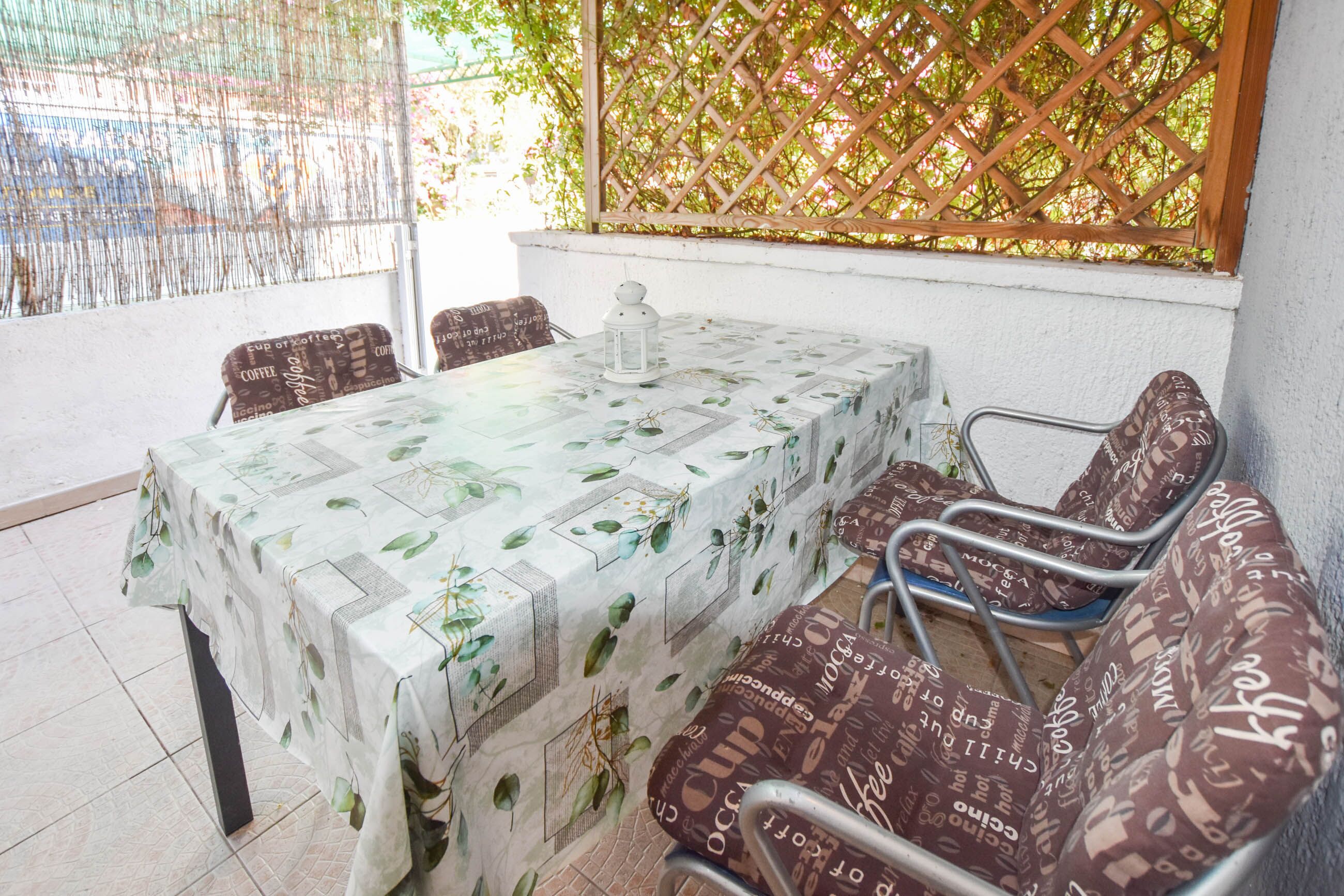 A dining area with a table covered in a leaf-patterned tablecloth, surrounded by metal chairs with patterned cushions.