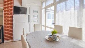 Un comedor con una mesa redonda cubierta por un mantel de lunares, cuatro sillas, una planta pequeña sobre una bandeja y una zona de cocina con un refrigerador y un televisor.