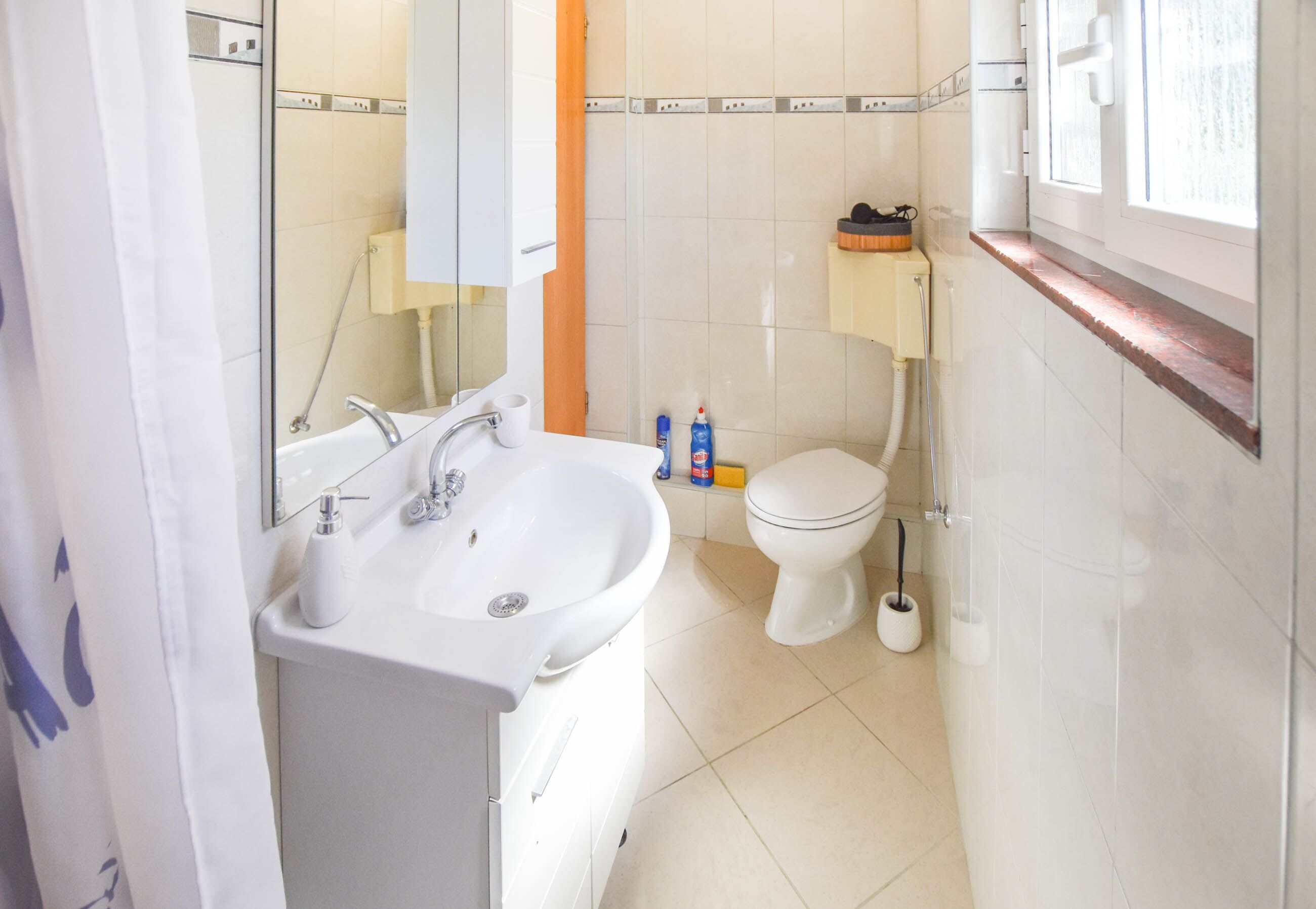 A bathroom with a white sink, mirror, and toilet. There are toiletries on the sink and a window above the toilet.