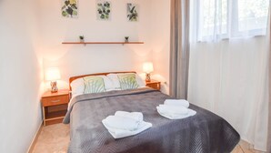 Una cama bien hecha con un cubrecolcha gris y dos toallas enrolladas, dos lámparas de noche, un pequeño estante con plantas y una ventana con cortinas traslúcidas.