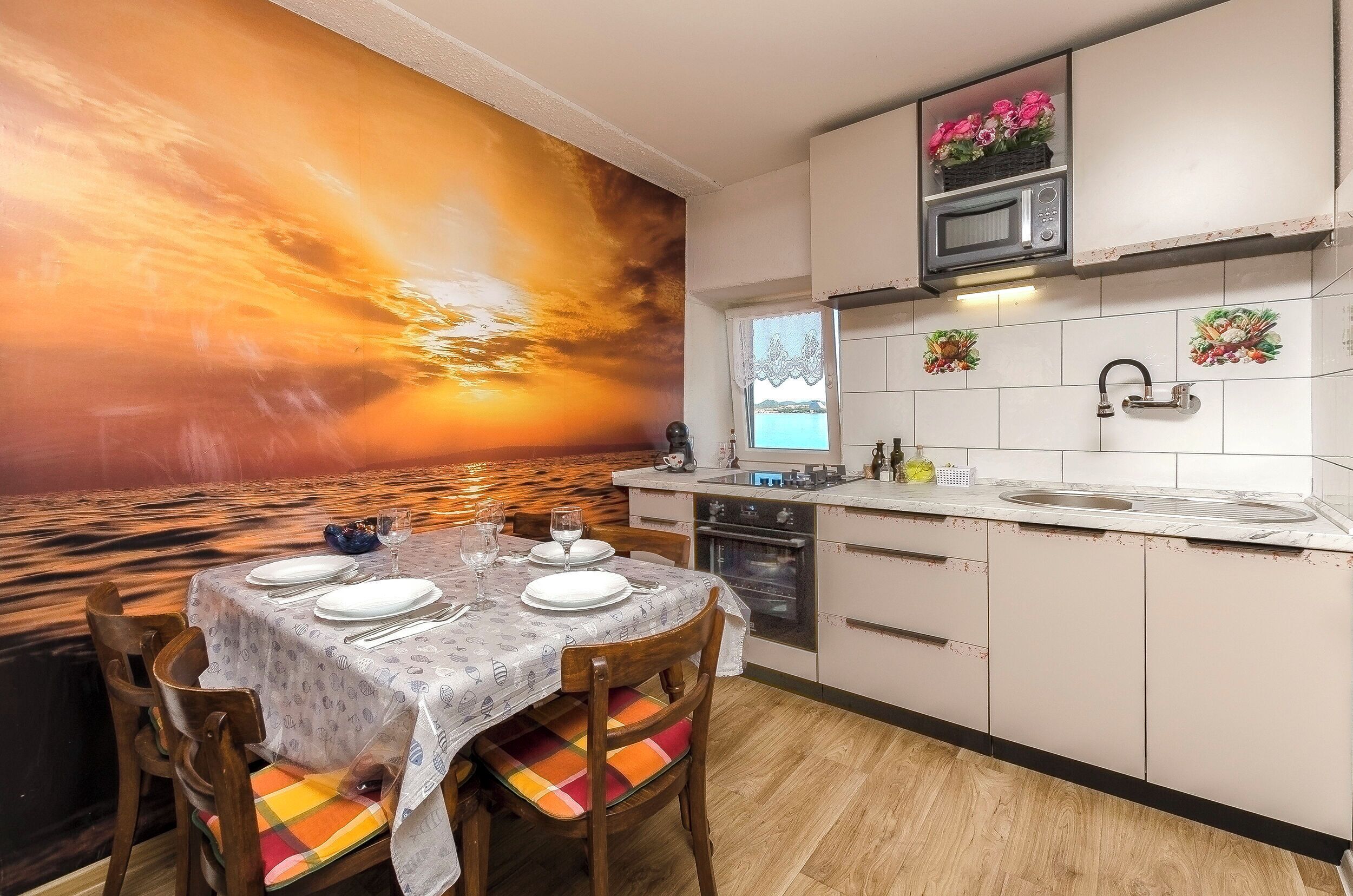 A dining area with a table set for four, a kitchen with white cabinets, a microwave, and a view of the sea through a window.
