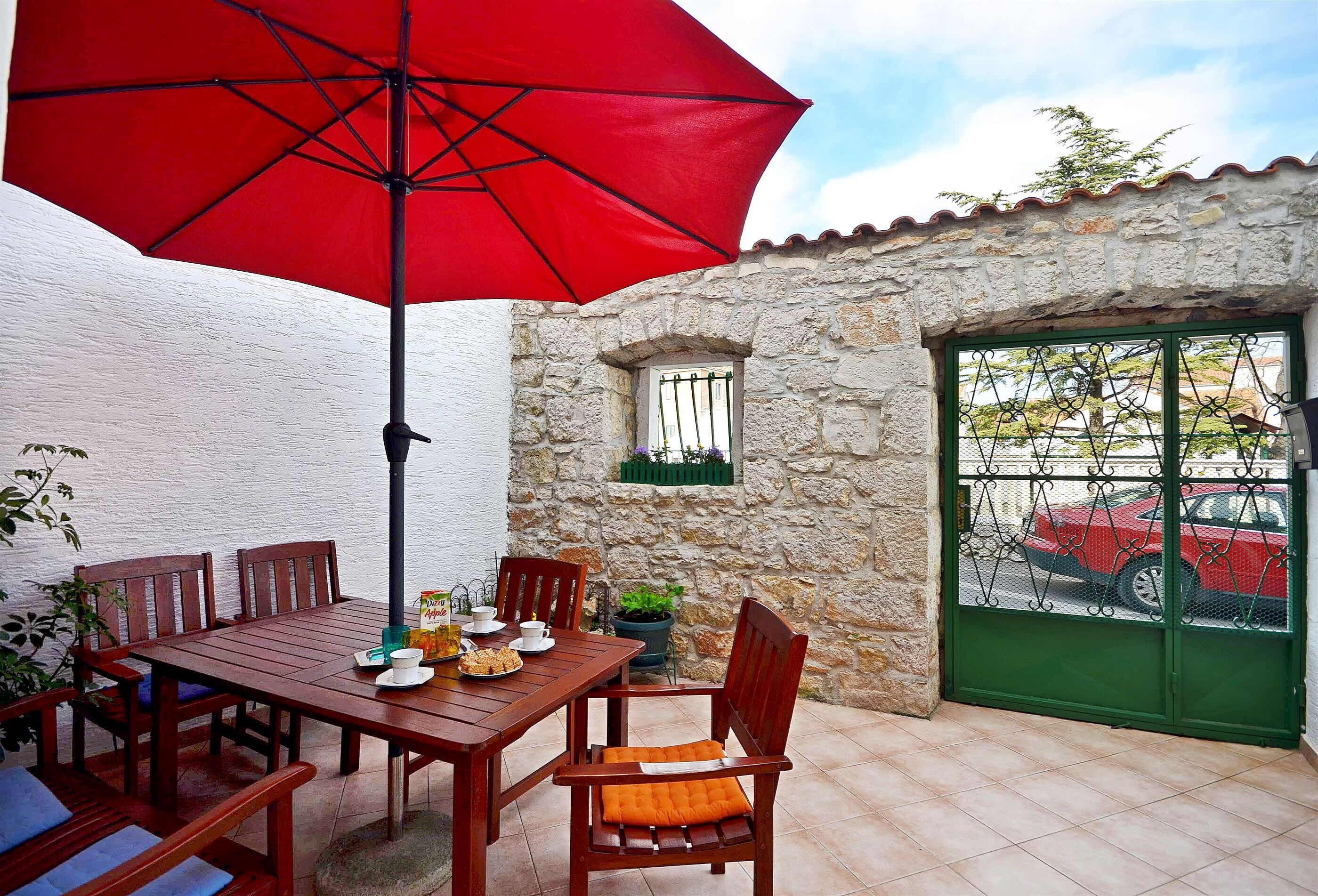 Uma mesa de madeira com cadeiras, um guarda-sol vermelho e uma parede de pedra com janela e porta.