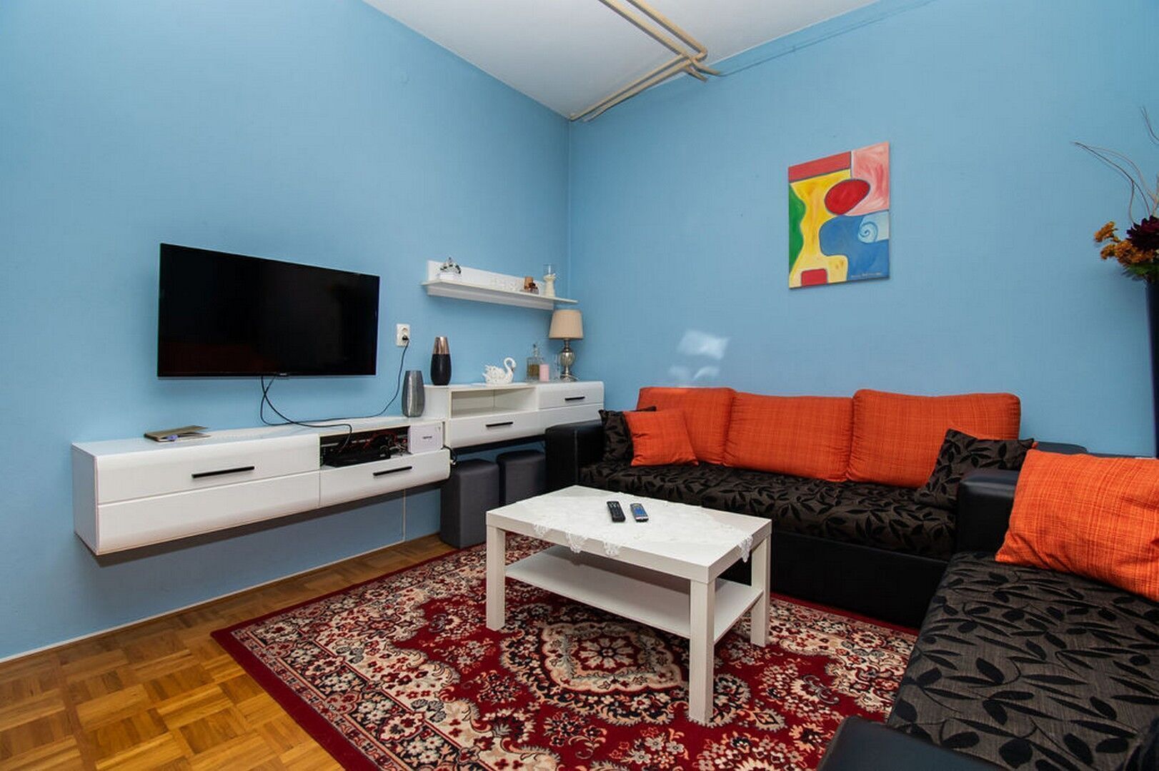 A living room with a flat-screen TV, a white coffee table, a black sofa with orange cushions, and a patterned rug.