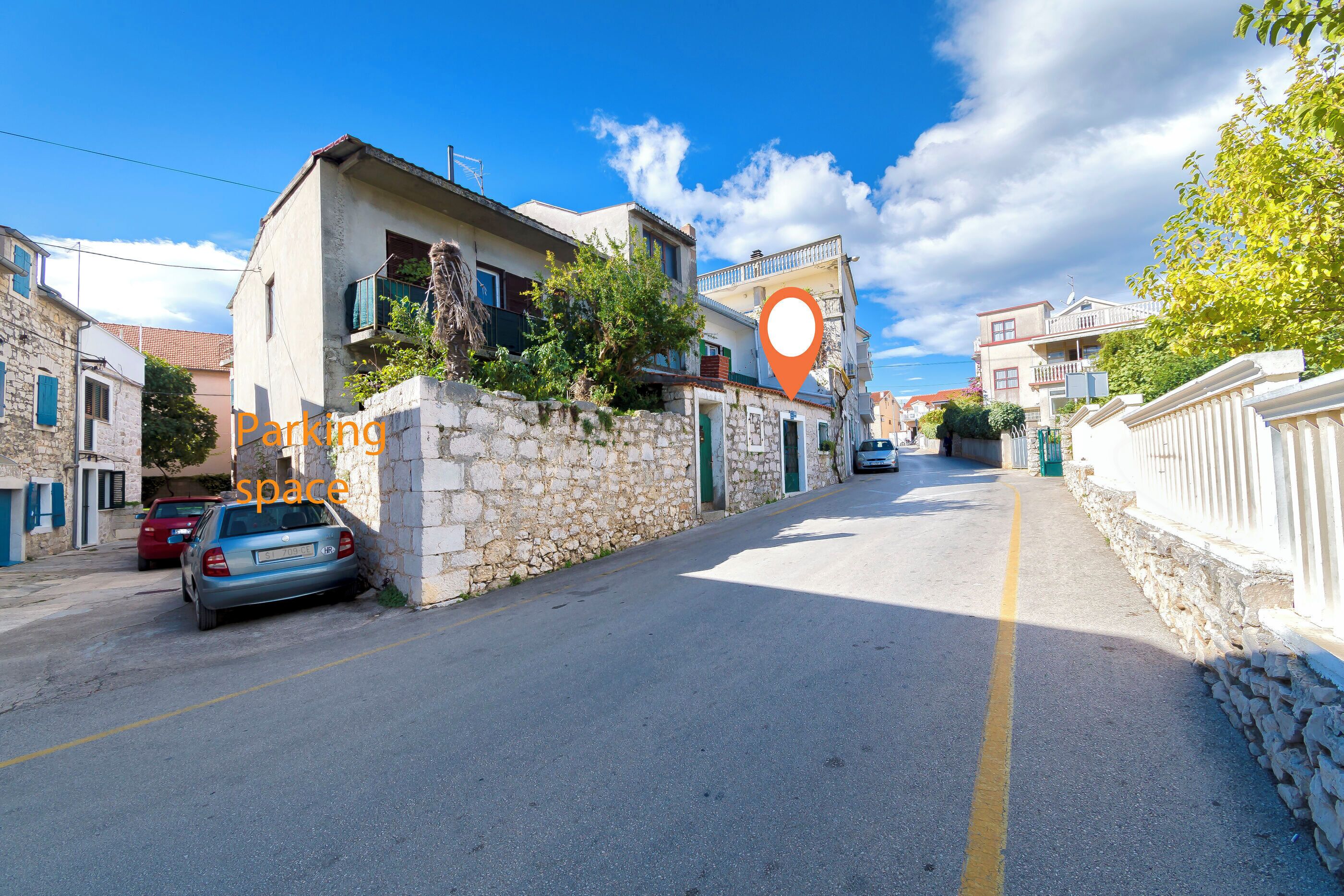 Una calle estrecha con muros de piedra y edificios, un coche estacionado, y un marcador de ubicación.