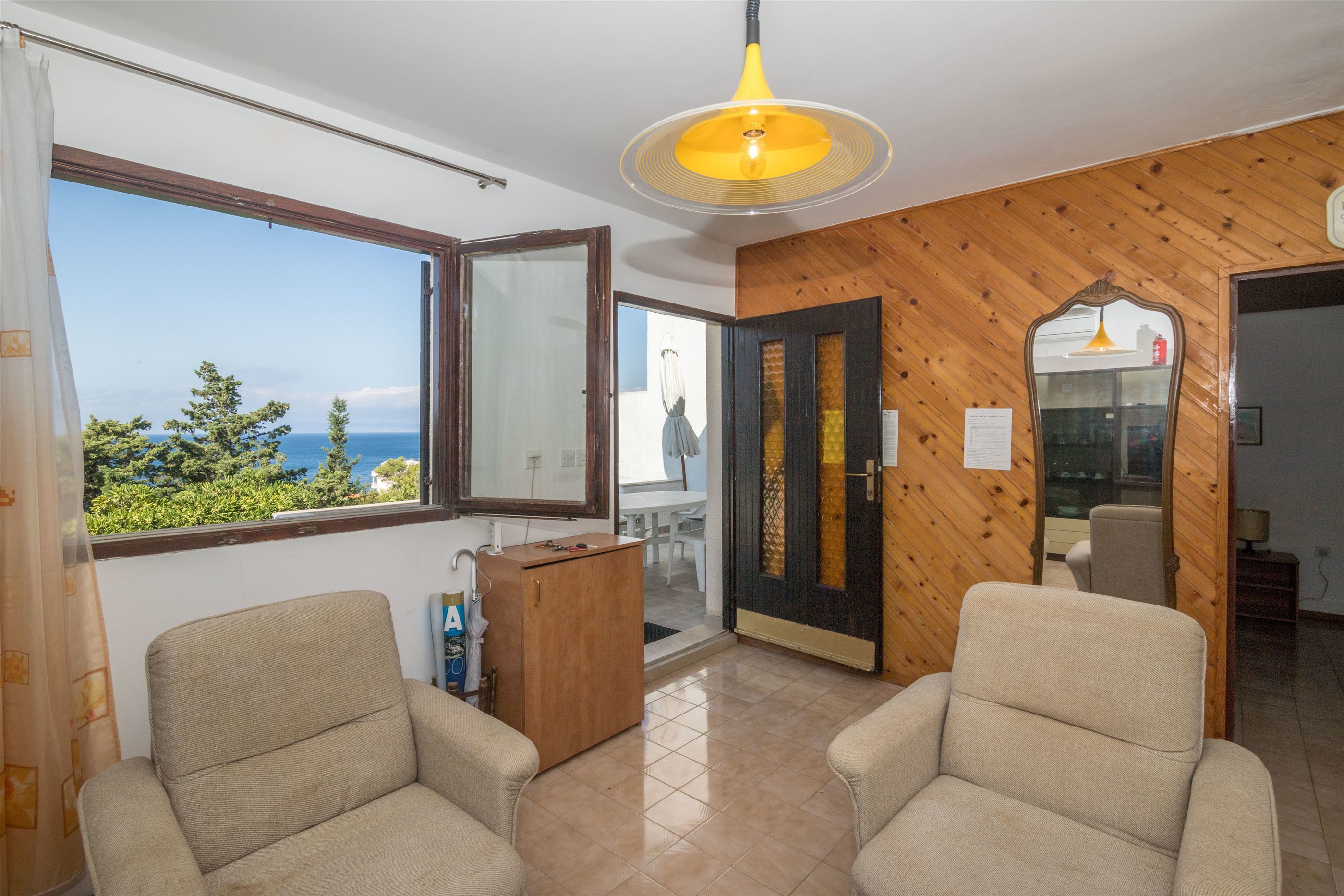 A room with two beige armchairs, a wooden cabinet, a large window with a view of the sea, and a yellow pendant light.