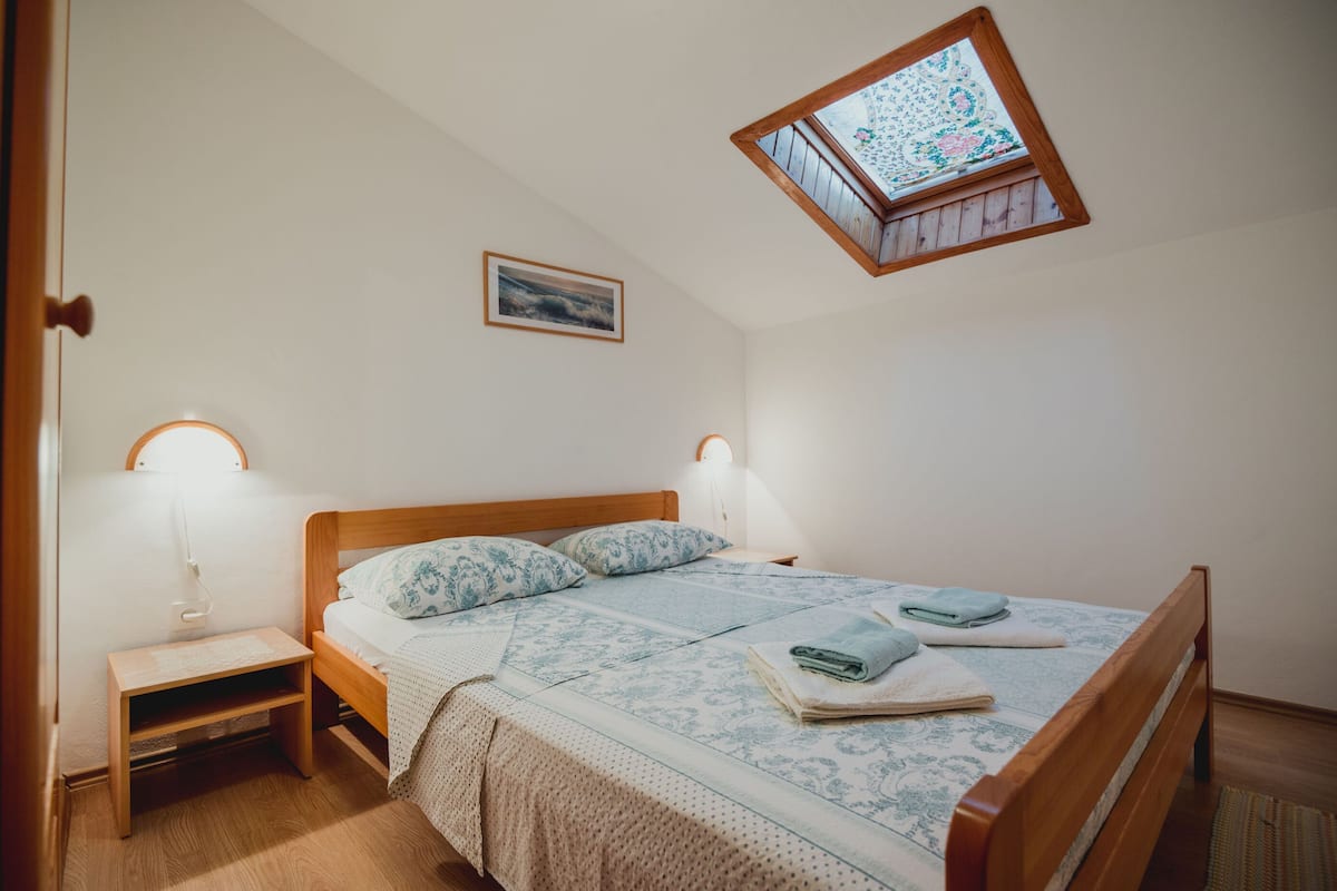 Un dormitorio con cama de madera, dos lámparas de noche, una mesita auxiliar pequeña, una ventana con vistas y un cuadro en la pared.