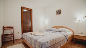 A bedroom with a wooden bed, a nightstand, a chair, a wooden door, and a wall-mounted light fixture. - Apartments Antun (Ivan Dolac)