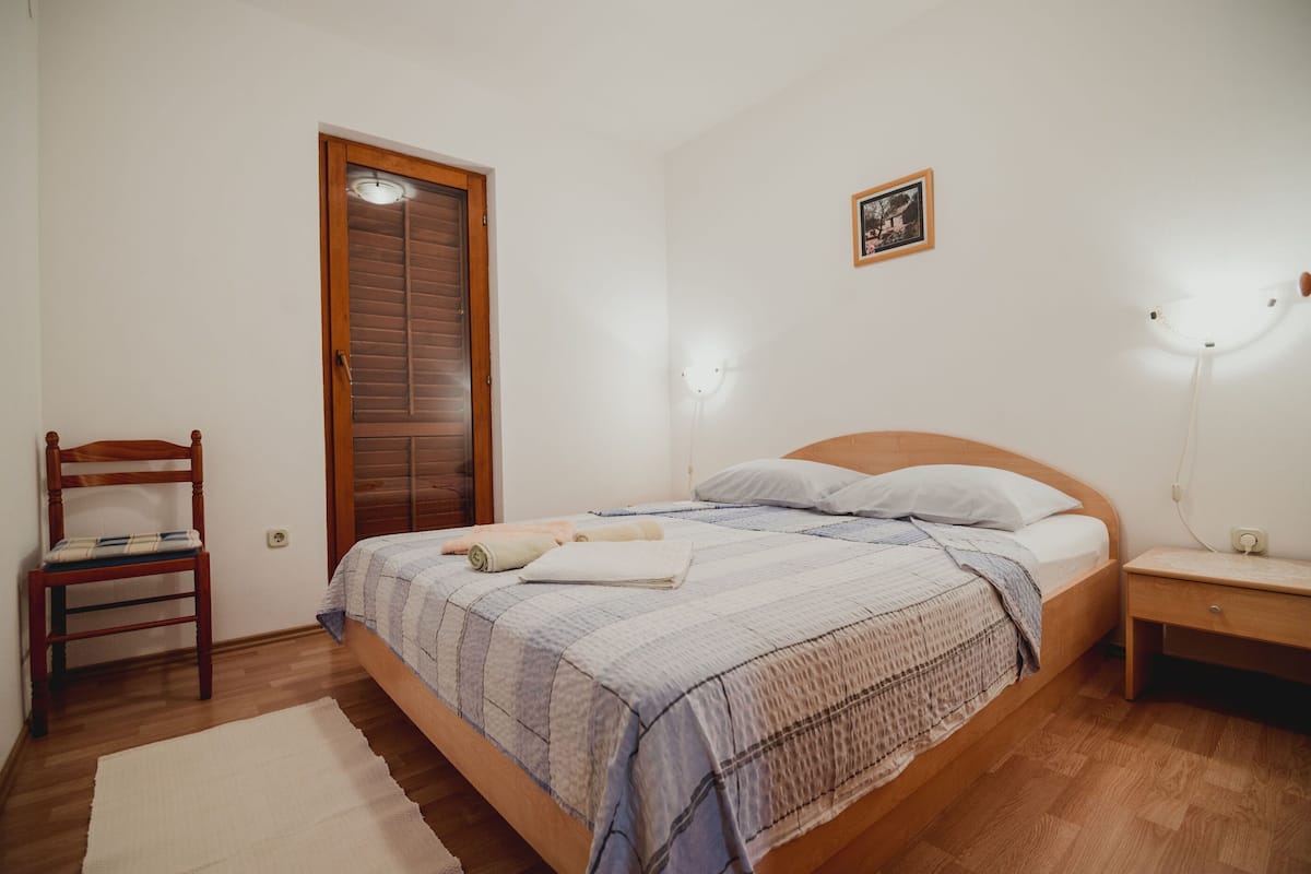 Un dormitorio con una cama de madera, una mesita de noche, una silla, una puerta de madera y una lámpara fijada en la pared.